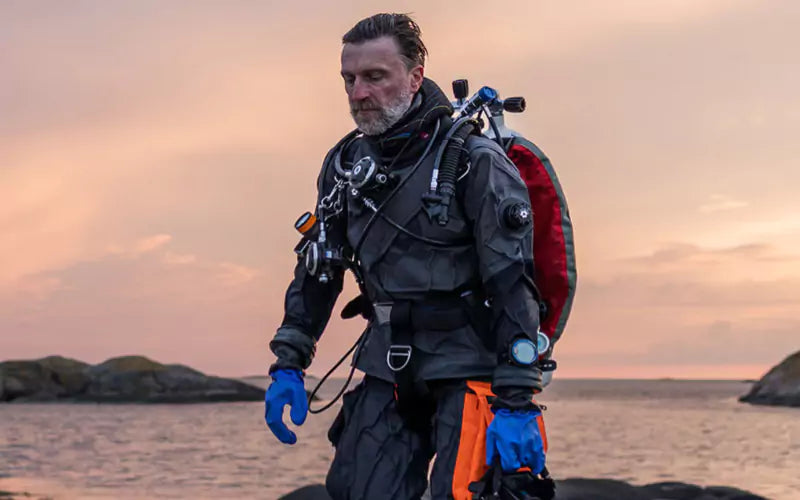 Man in Avatar drysuit walking on rocks with a sunset sea in the background